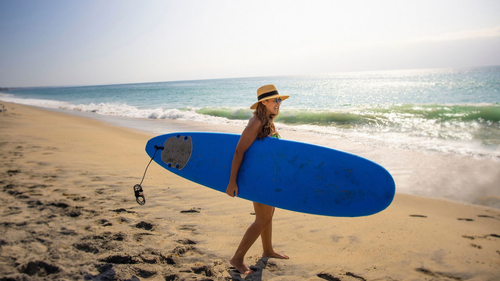 Frau mit Surfboard am Strand mit Hut und Sonnnebrille