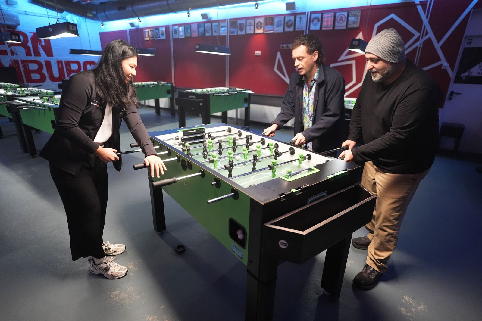 Tischkicker-Weltmeisterin Linh Tran zeigt ihr Können den beiden Schauspielern.