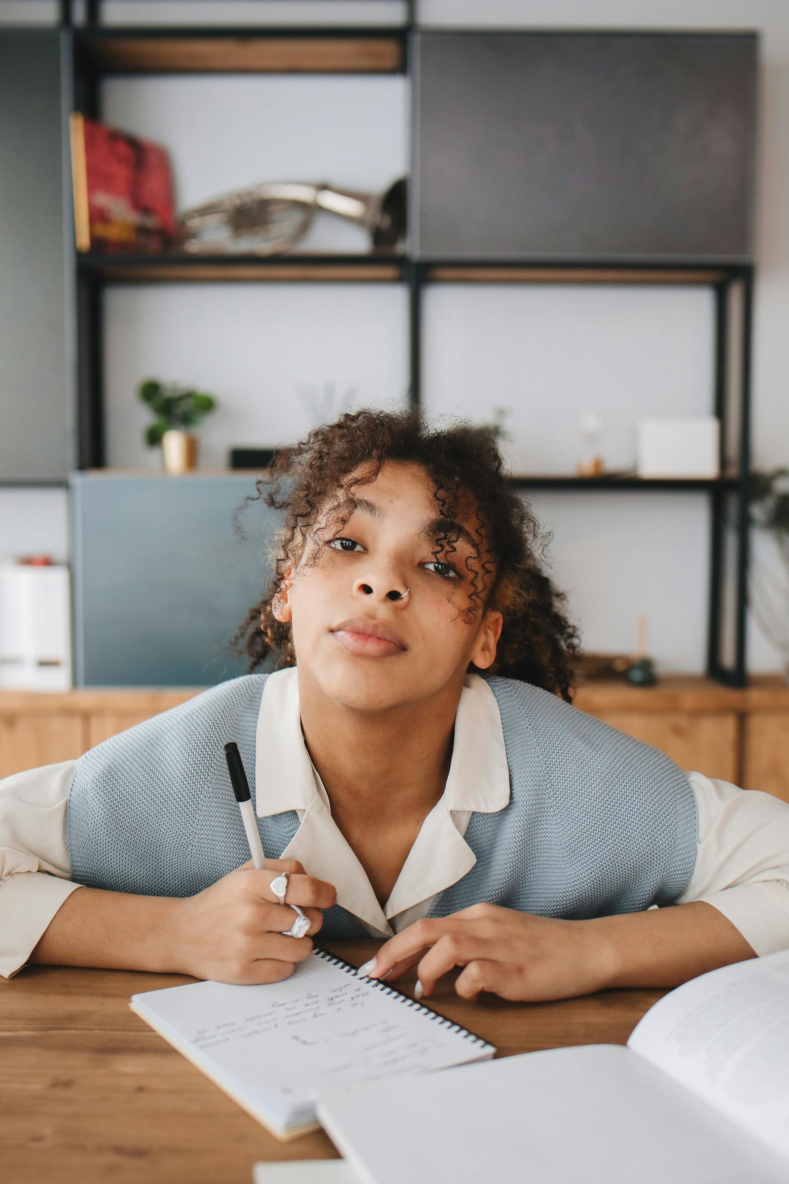 Frau mit Locke sitzt an Schreibtisch und schaut in die Kamera und hält Stift in der Hand, sie ist auf Tisch gelehnt