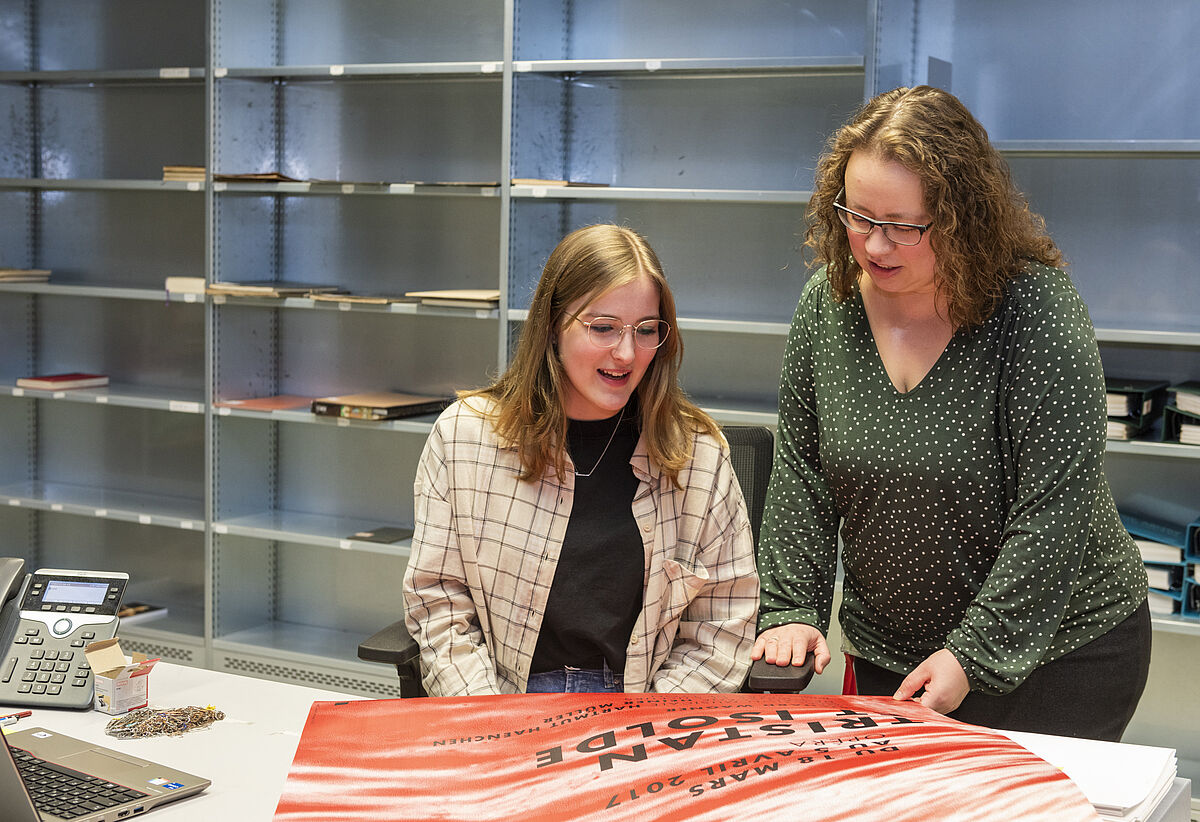 Zwei Frauen stehen neben einander an einem Tisch und schauen auf ein Plakat