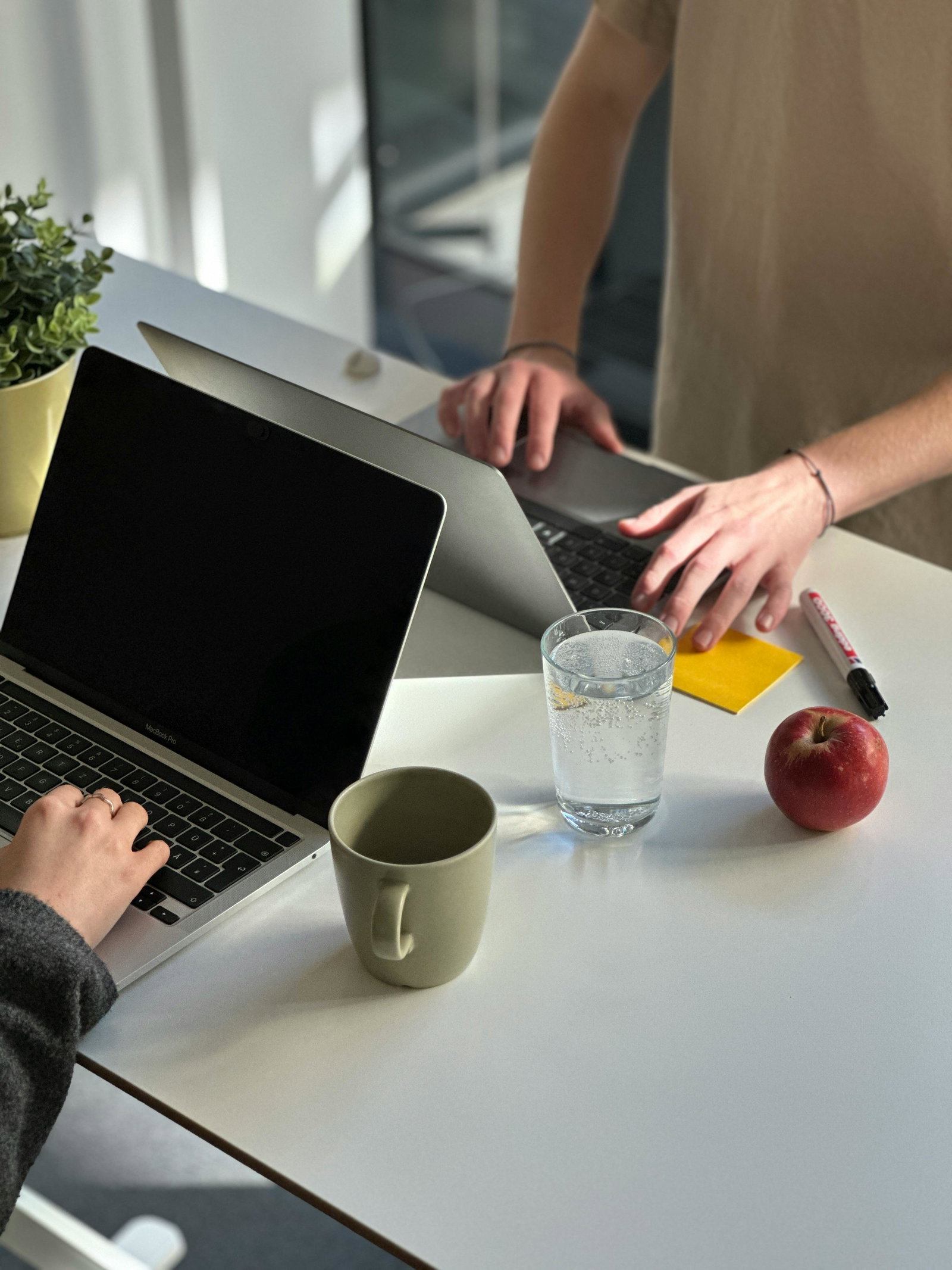 Zwei Personen stehen mit Laptop gegen über und tippen auf Tisch mit Wassergläsern und einem Apfel