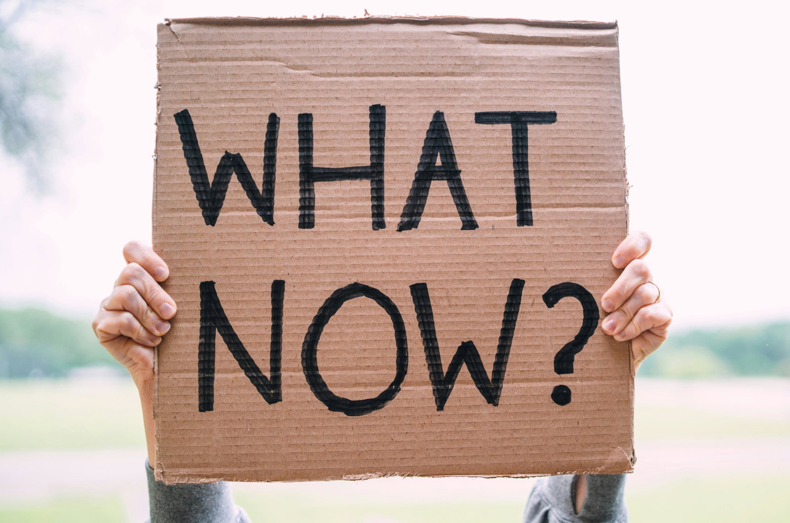 Hände heben ein Pape-Schild mit Text "What Now?" hoch