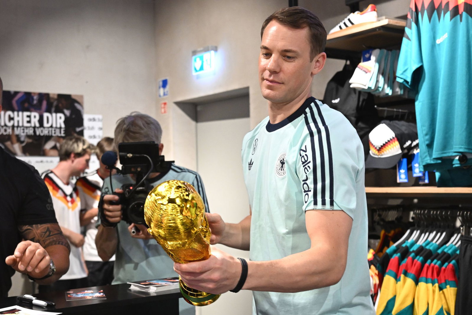 Lächelnd signiert Manuel Neuer einen goldenen WM-Pokal. 