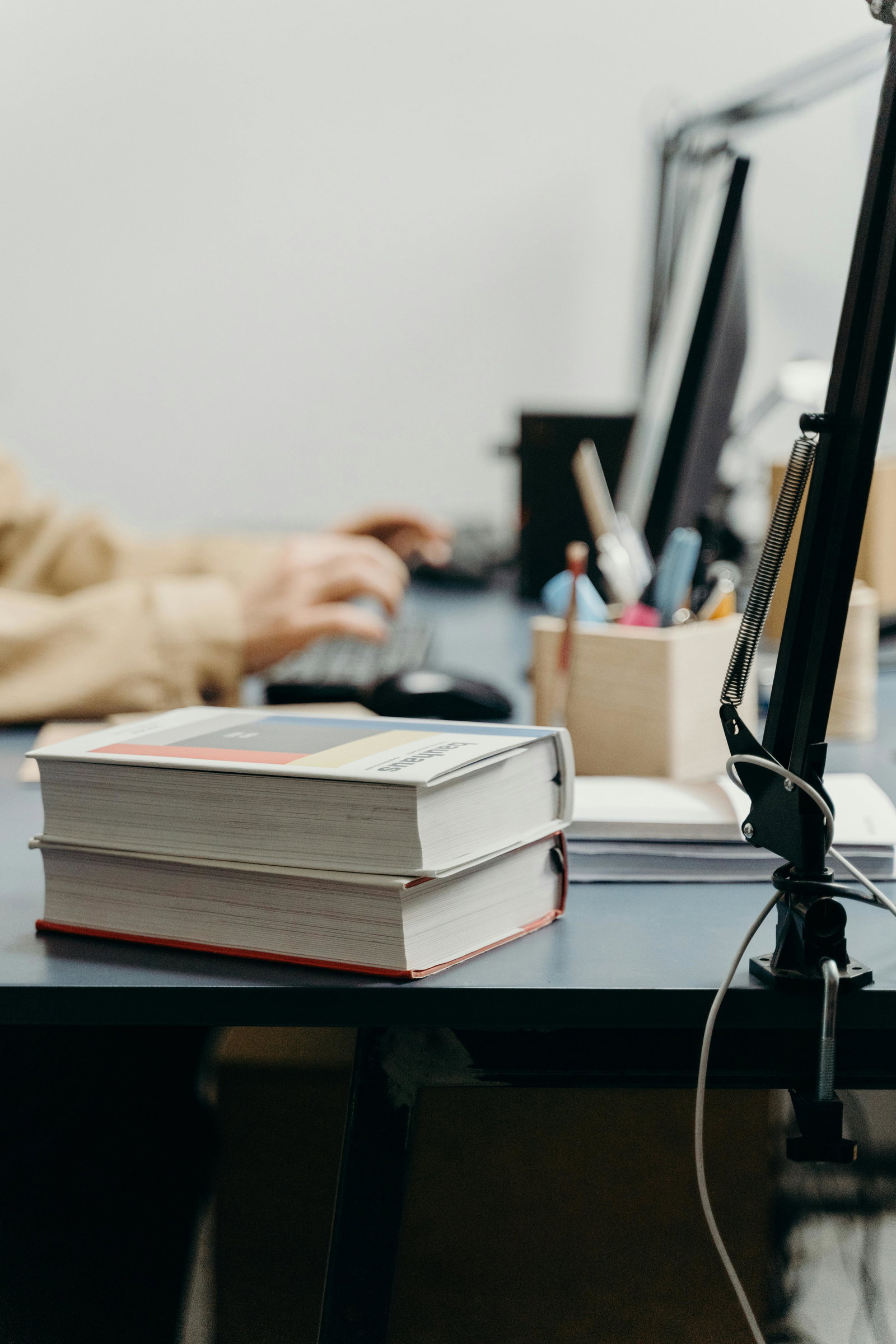 Bücher liegen im Vordergrund auf Schreibtisch, im Hintergrund sieht man verschwommen eine Person die am Laptop arbeitet