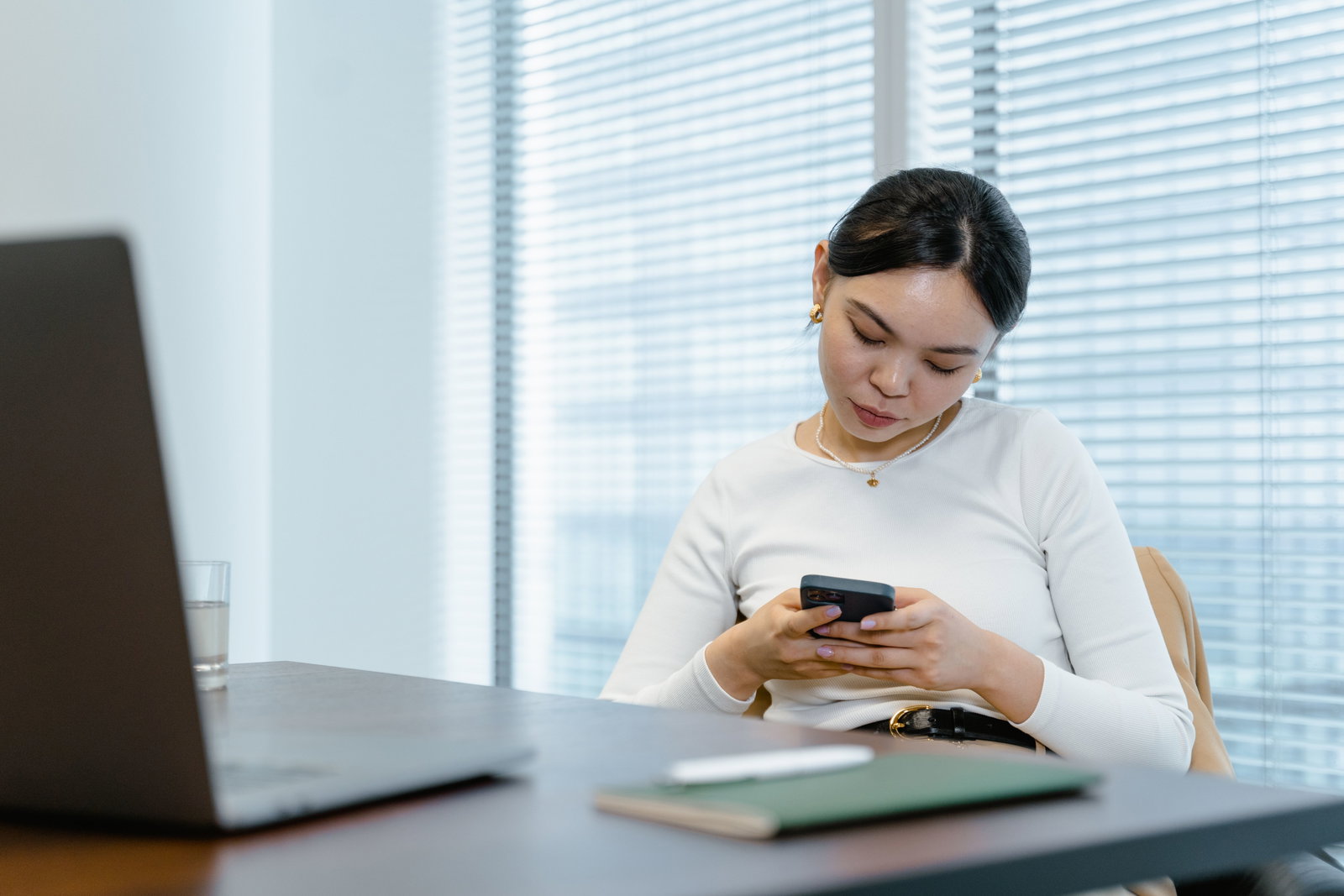 Sitzende Frau am Tisch, wo ein Laptop, Wasserglas und Heft zu sehen ist während sie auf ihr Handy schaut 
