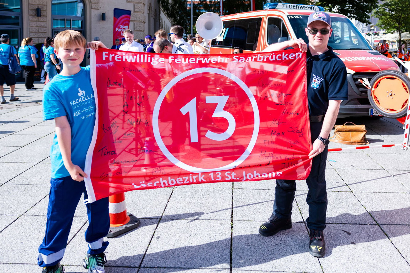 Ein Jugendfeuerwehrmann (l.) und ein aktiver Feuerwehrmann der Freiwilligen Feuerwehr Saarbrücken-St. Johann (LB 13) präsentieren ein besonderes Banner mit der Zahl 13.
