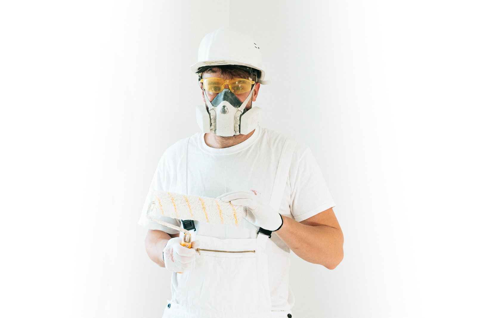 Maler und Lackiere mit Schutzmaske und Helm schaut in die Kamera und hält eine Farbwalze