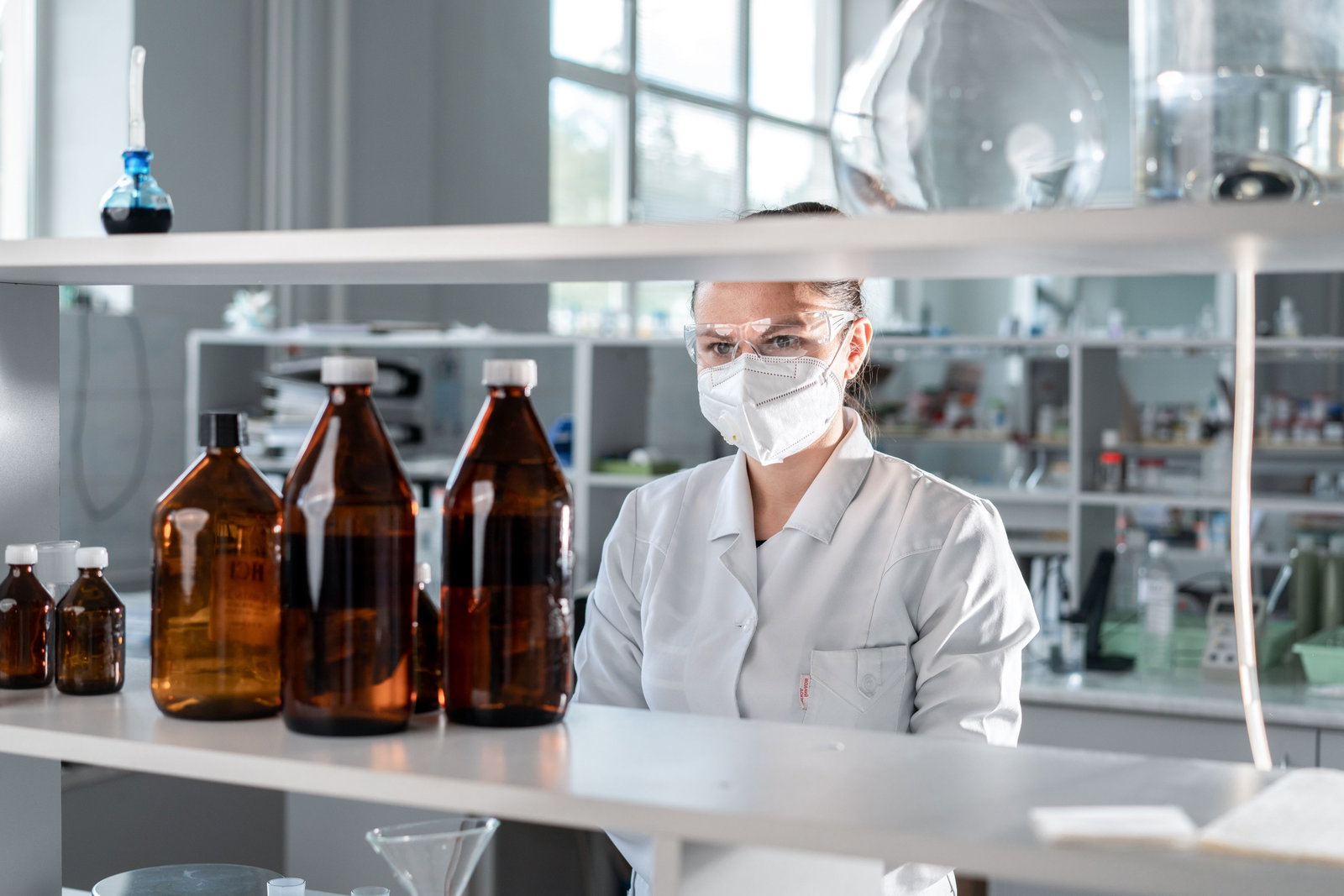 Pharmakantin steht mit Laborkitttel, Schutzbrille und Maske vor großen Laborflaschen