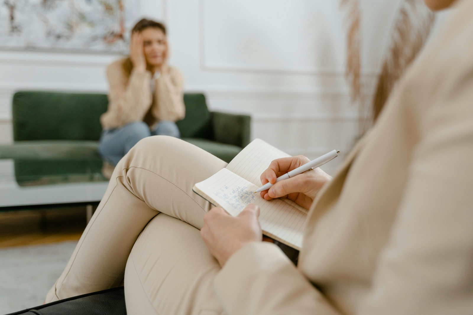 Frau sitzt im Hintergrund auf einem Sofa und schaut verzweifelt, im Vordergrund sitzet eine Frau auf einem Sesel und macht Notizen
