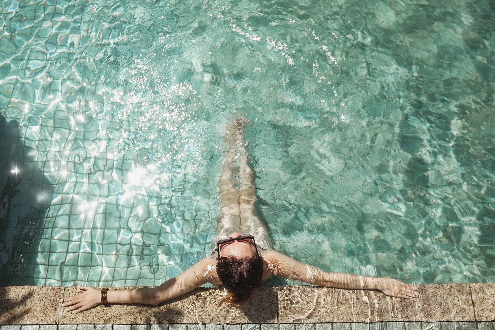 Frau sitzt im Pool mit ausgestreckten armen und Sonnenbrille