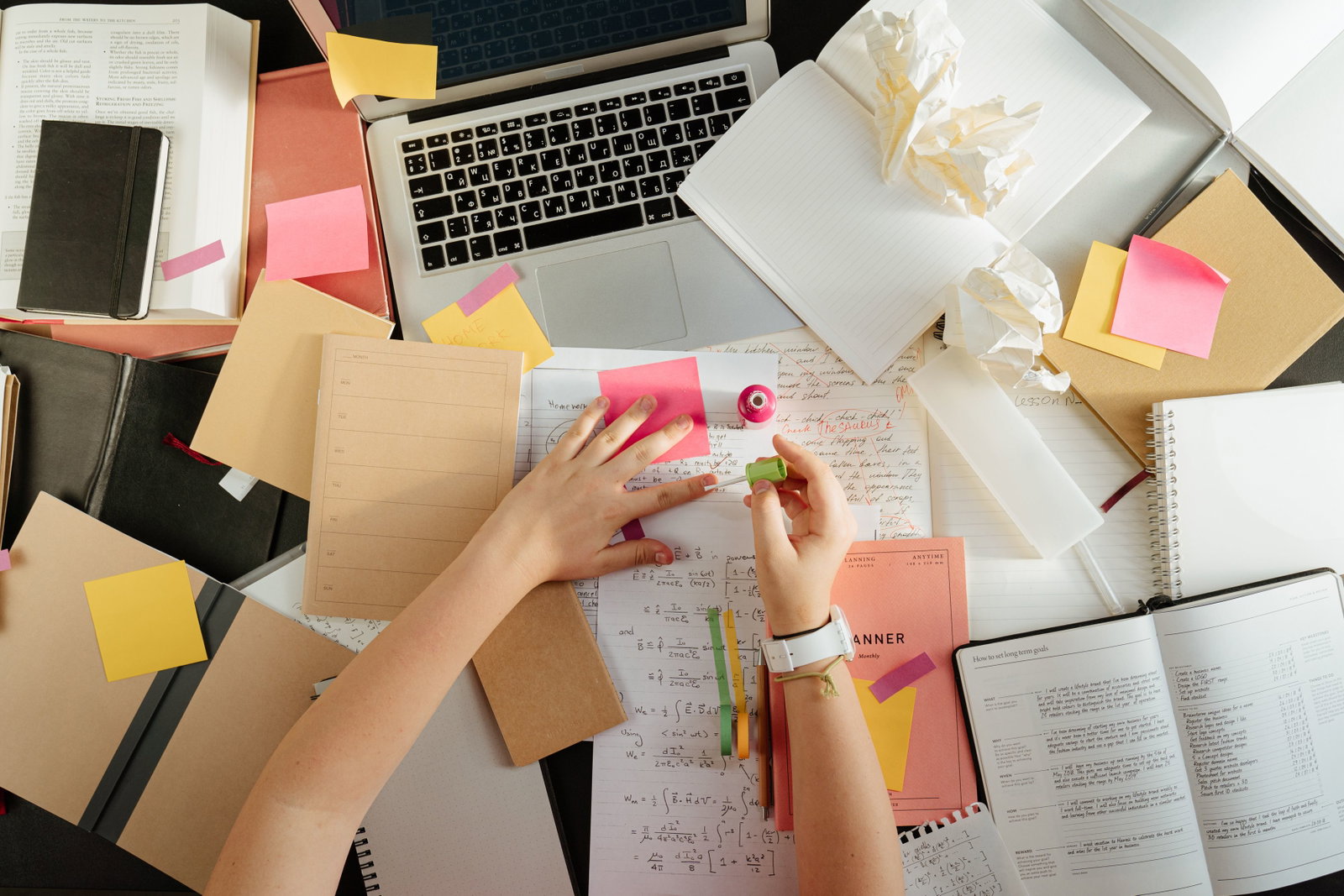 Unordentlicher Schreibtisch, mit Blättern, Laptop und Händen, die Fingernägel lackieren