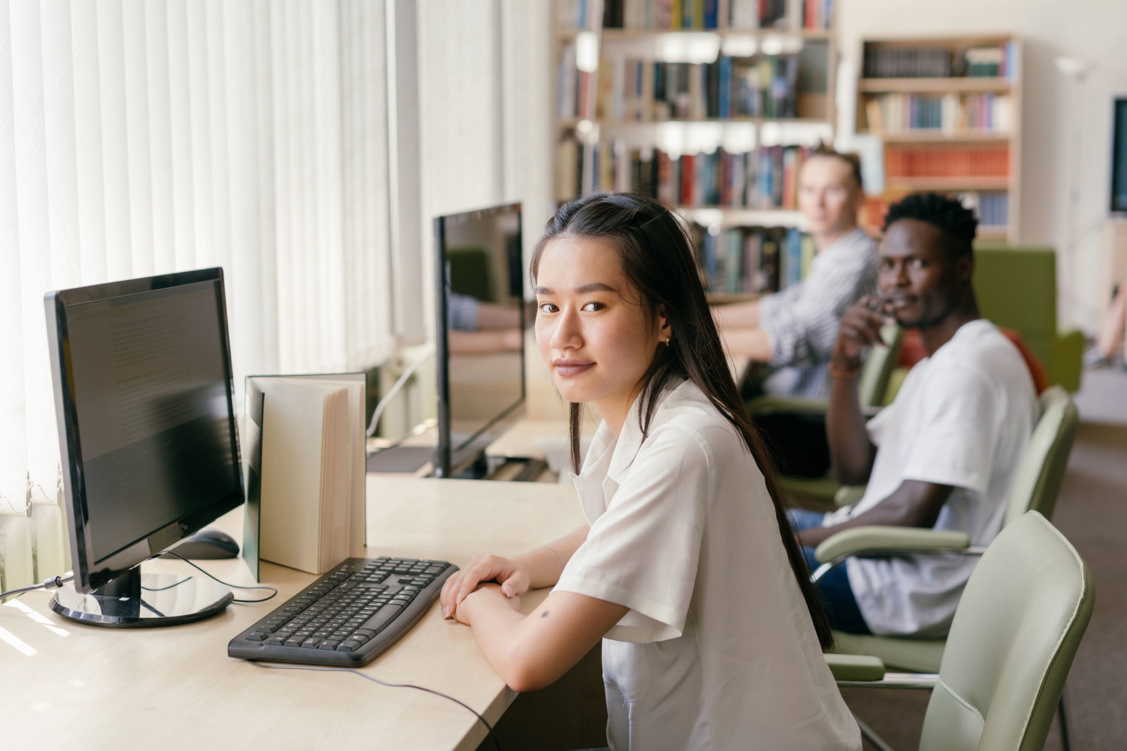 Drei Studenten (Mädchen und zwei Jungs) sitzen vor Computer am Schreibtisch und schauen in die Kamera