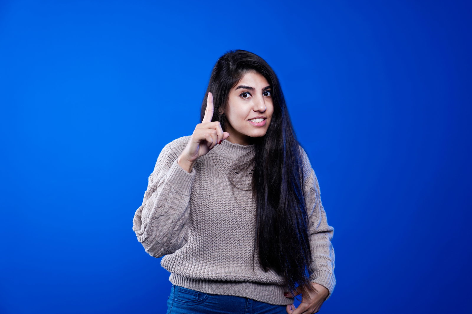 Frau mit dunkle n Haaren trägt einen Pullover und steht vor einem blauen Hintergrund und hält einen Finger hoch