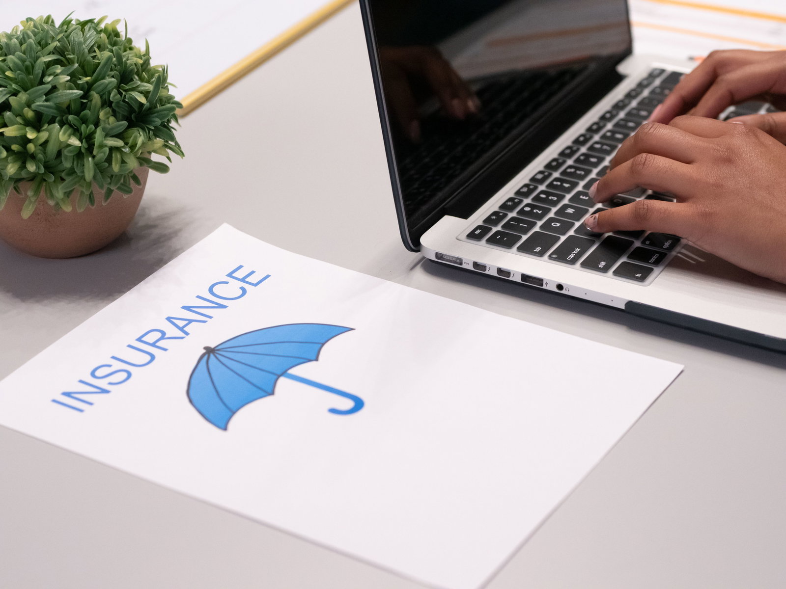 Männer Hände tippen auf PC, auf dem Tisch daneben liegt ein Blatt mit der Aufschrift INSURANCE und einem blauen Regenschirm darauf abgebildet
