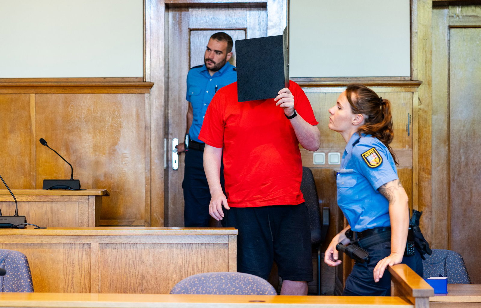 Justizvollzugsbeamte führen den Angeklagten, der sein Gesicht mit einem Aktenordner verdeckt, vor Beginn der Verhandlung in den Saal 137 des Landgerichts.