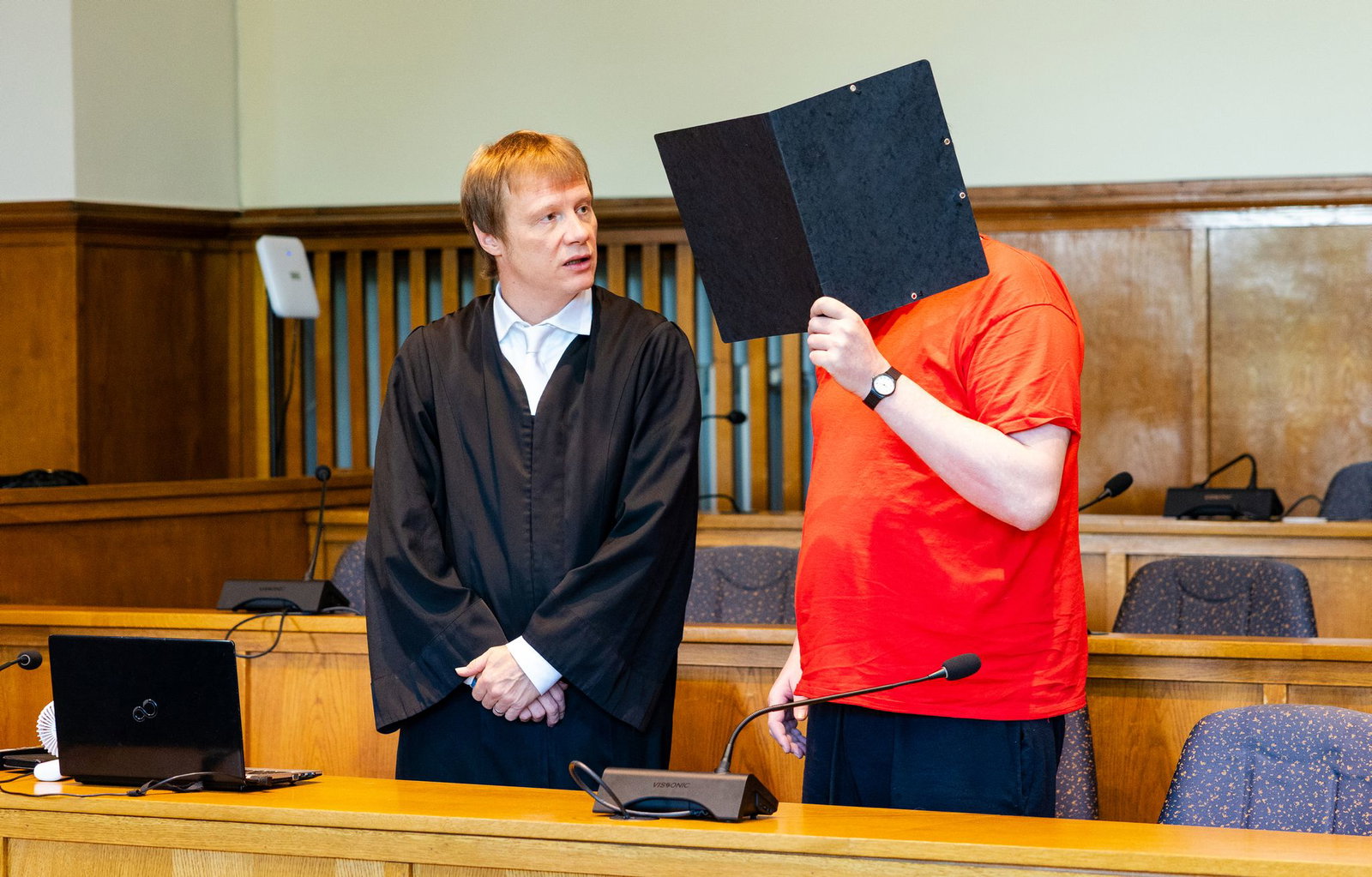 Der Angeklagte (r) steht mit seinem Verteidiger Dirk Gerlach am Verhandlungstisch im Saal 137 des Landgerichts und verdeckt sein Gesicht mit einem Aktenordner.
