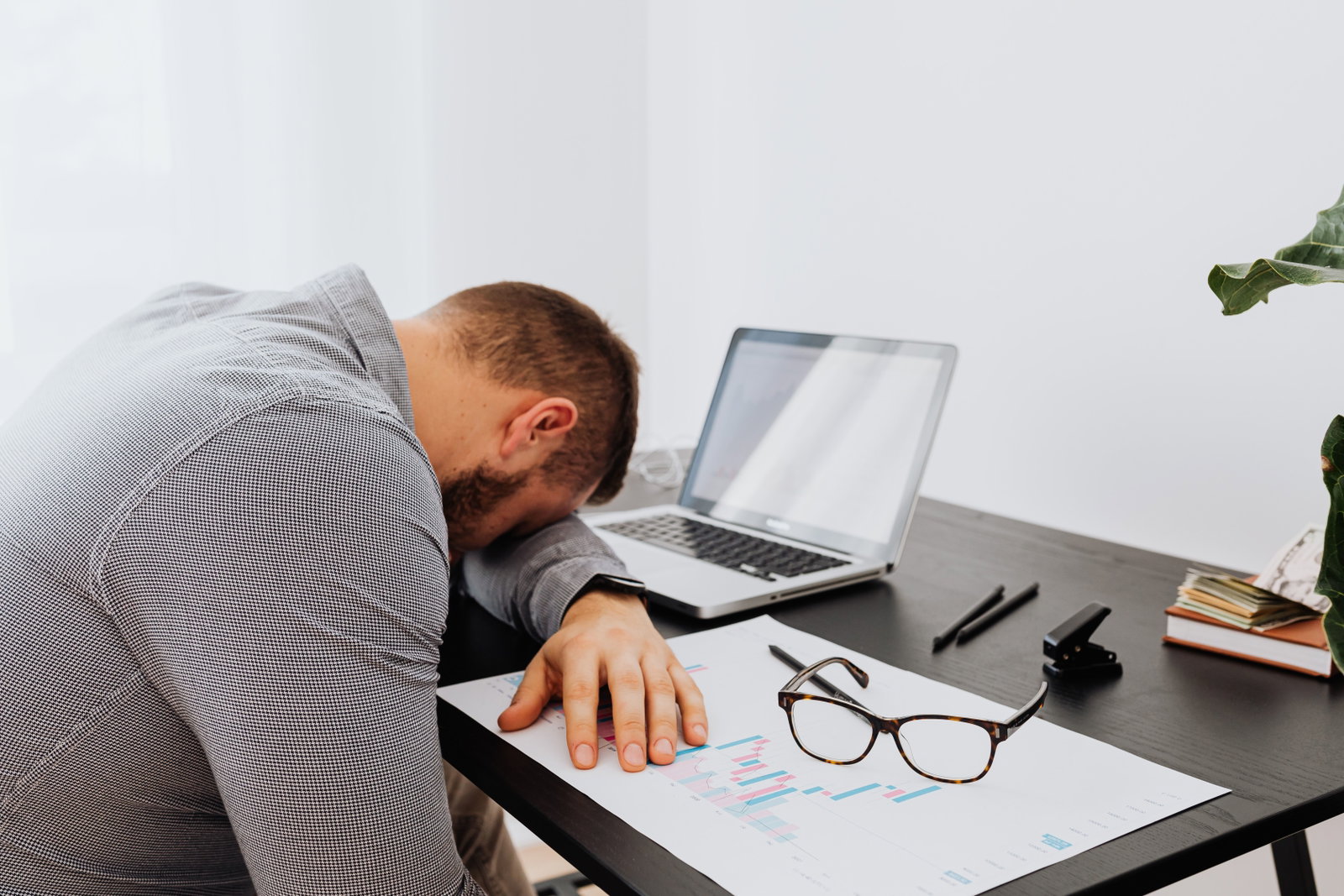Ein Mann mit einem grauen Hemd sitzt müde an einem Schreibtisch mit einem Laptop und Dokumenten und zeigt Anzeichen von Stress am Arbeitsplatz.