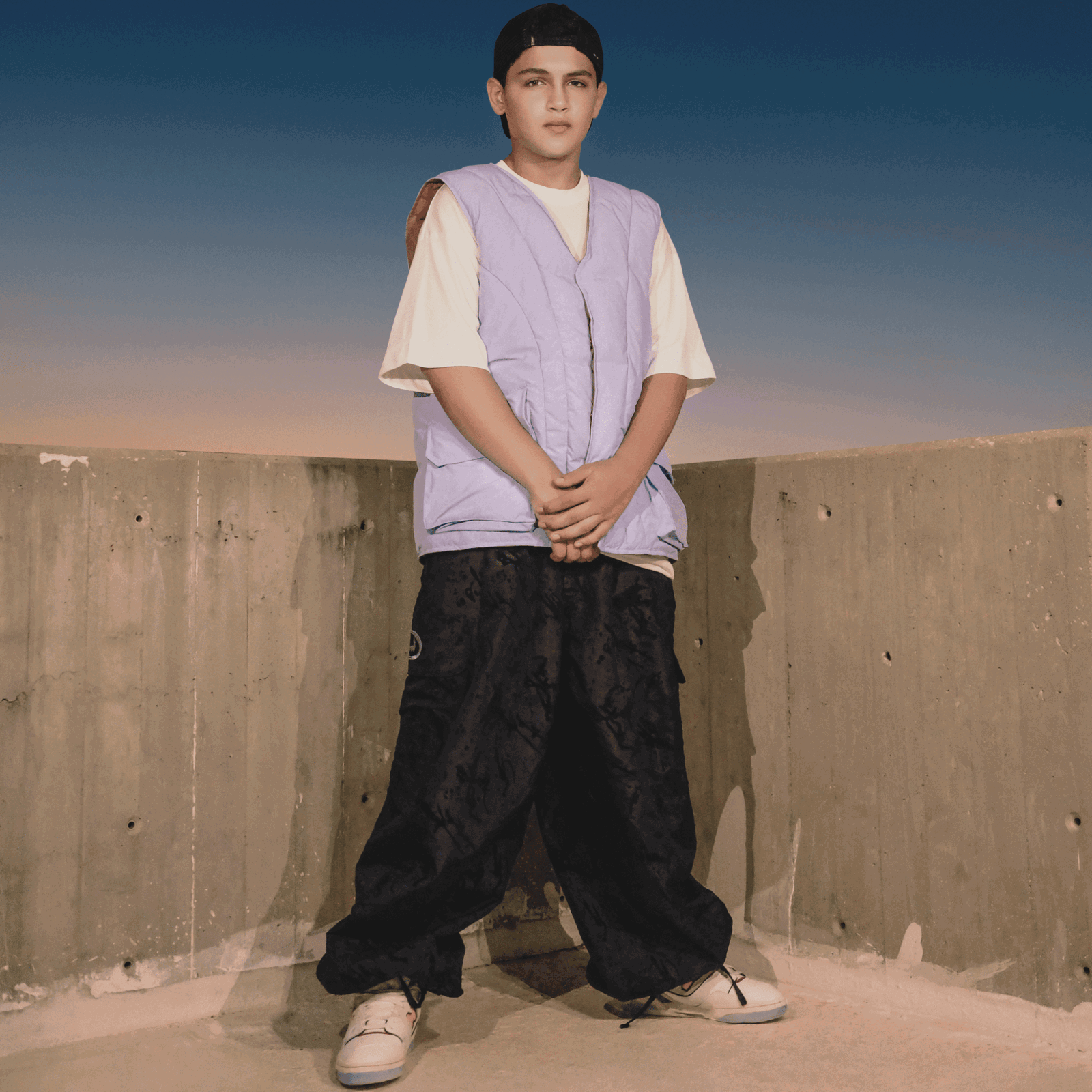 Der palästinensische Rapper MC Abdul steht vor einer Betonwand im Abendlicht. Er trägt eine blaue Weste, weite schwarze Hosen, Sneaker und eine rückwärts getragene Cap. Sein Blick ist ruhig, die Stimmung nachdenklich.