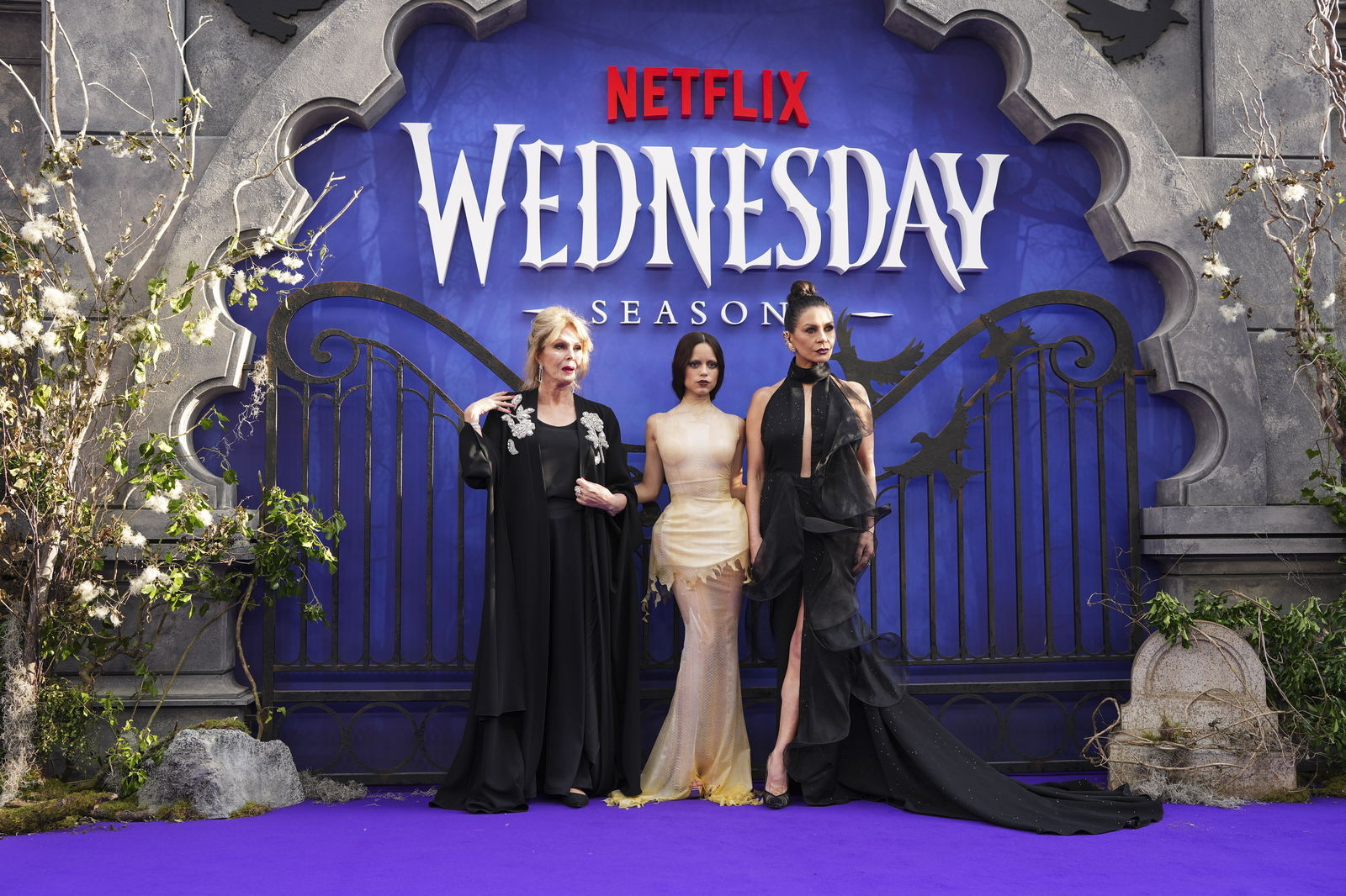 Joanna Lumley, Jenna Ortega und Catherine Zeta-Jones posieren elegant auf einem lila Teppich vor einem düsteren Tor-Backdrop mit dem Schriftzug Netflix Wednesday Season 2.